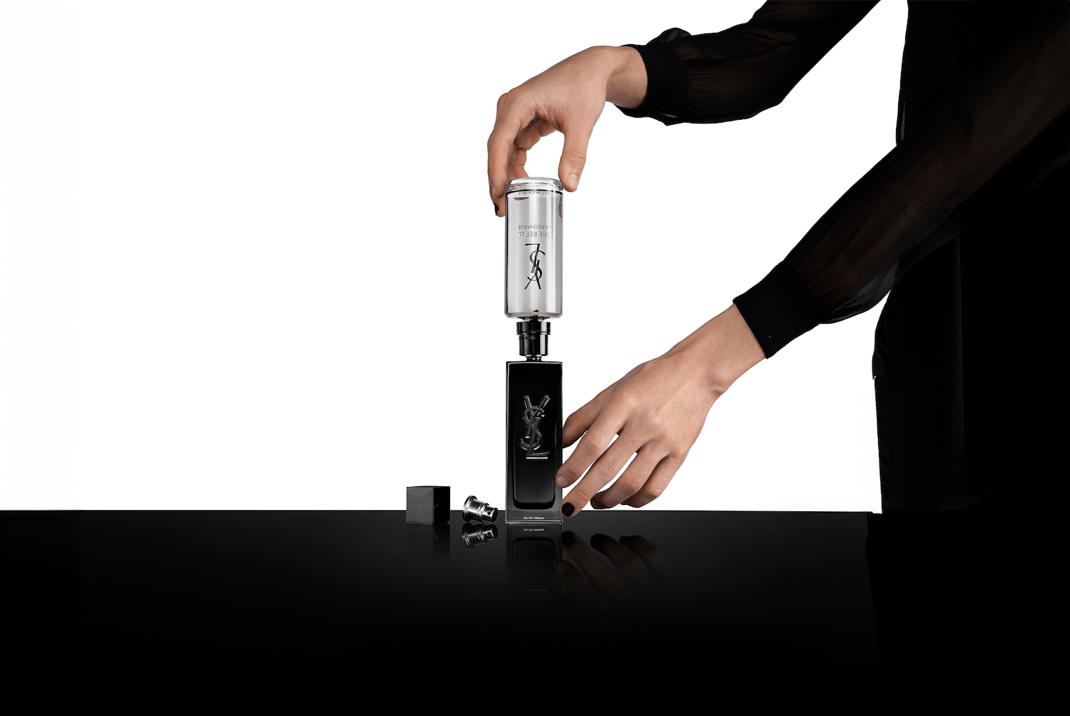 Eine Person in schwarzer Kleidung füllt eine schwarze YSL-Parfümflasche mit einer silbernen Nachfüllpackung auf einem glänzend schwarzen Tisch nach.