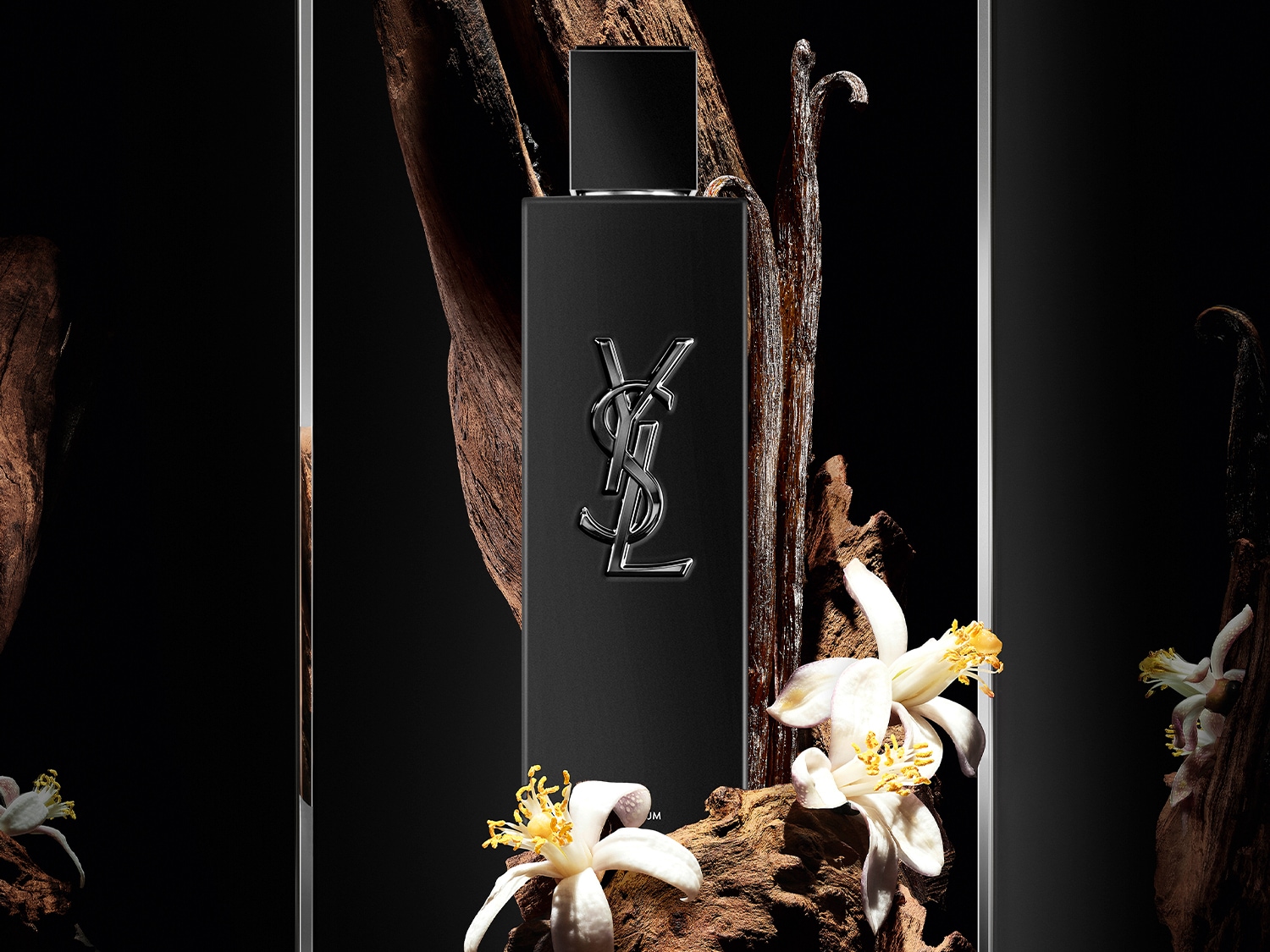 Schwarzer YSL-Flakon mit silbernem Logo, umgeben von Holzstücken und weißen Blüten.
