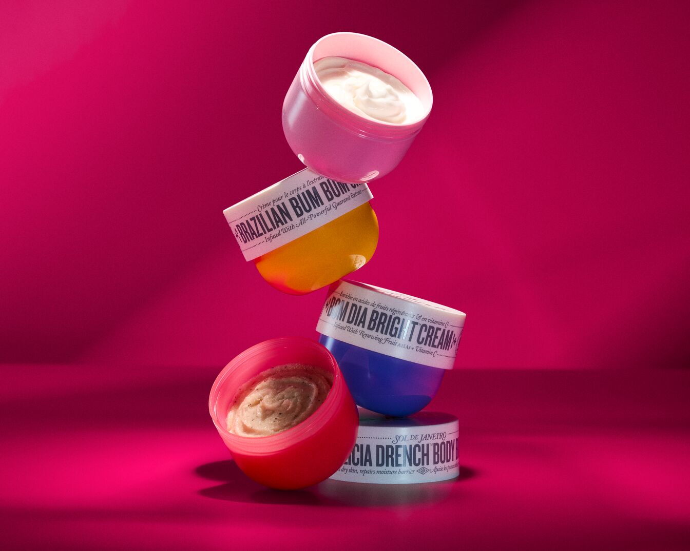 Auf einem pinkfarbenen Hintergrund sind gestapelte Sol de Janeiro Körpercremes in Rosa, Gelb, Blau und Rot zu sehen, darunter Brazilian Bum Bum, Bom Dia Bright Cream und Cia Drench Body.