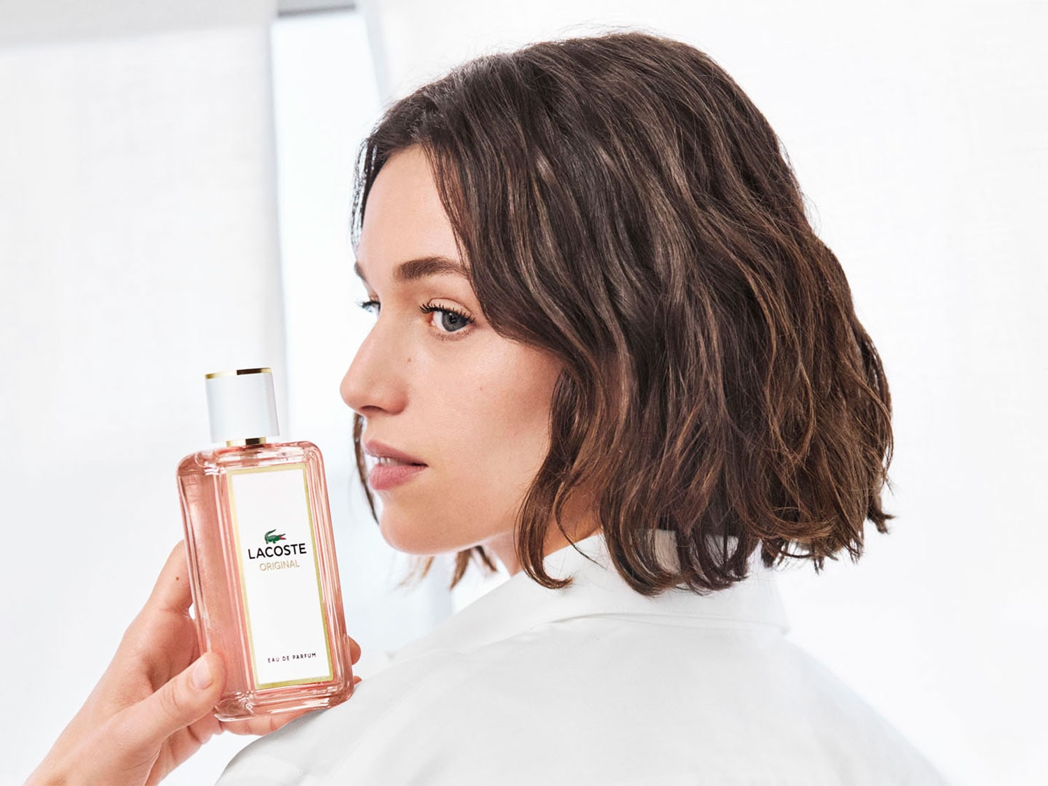 Kobieta trzyma w dłoni flakon Lacoste Original Eau de Parfum o pojemności 50 ml, z logo krokodyla na białej etykiecie i delikatnym różowym odcieniem płynu.