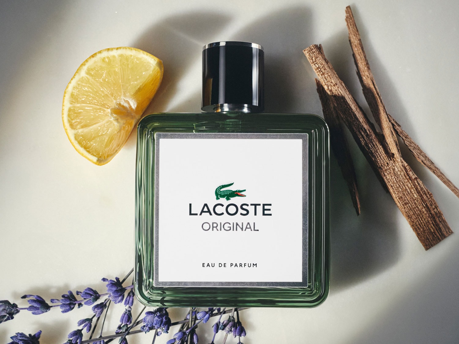 Butelka zielonego perfumu Lacoste Original Eau de Parfum z plasterkiem cytryny, lawendą i drewienkami.