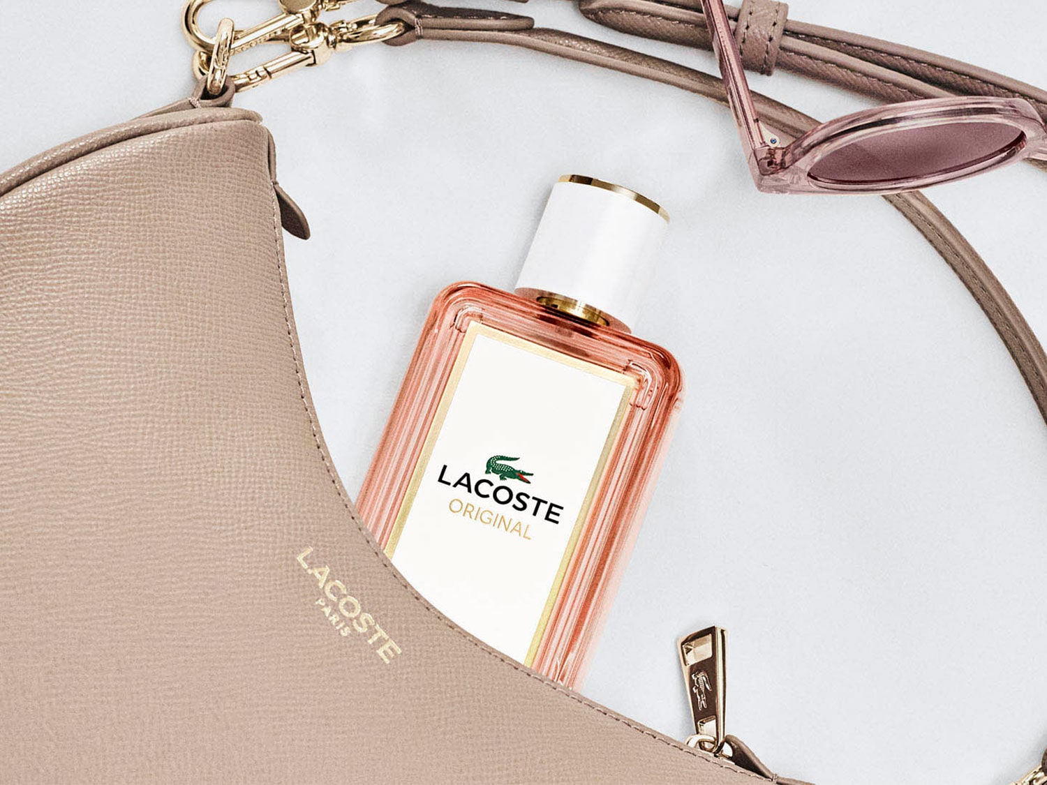 Flakon perfum Lacoste Original w otoczeniu beżowej torebki i okularów przeciwsłonecznych, tworzący elegancką kompozycję.