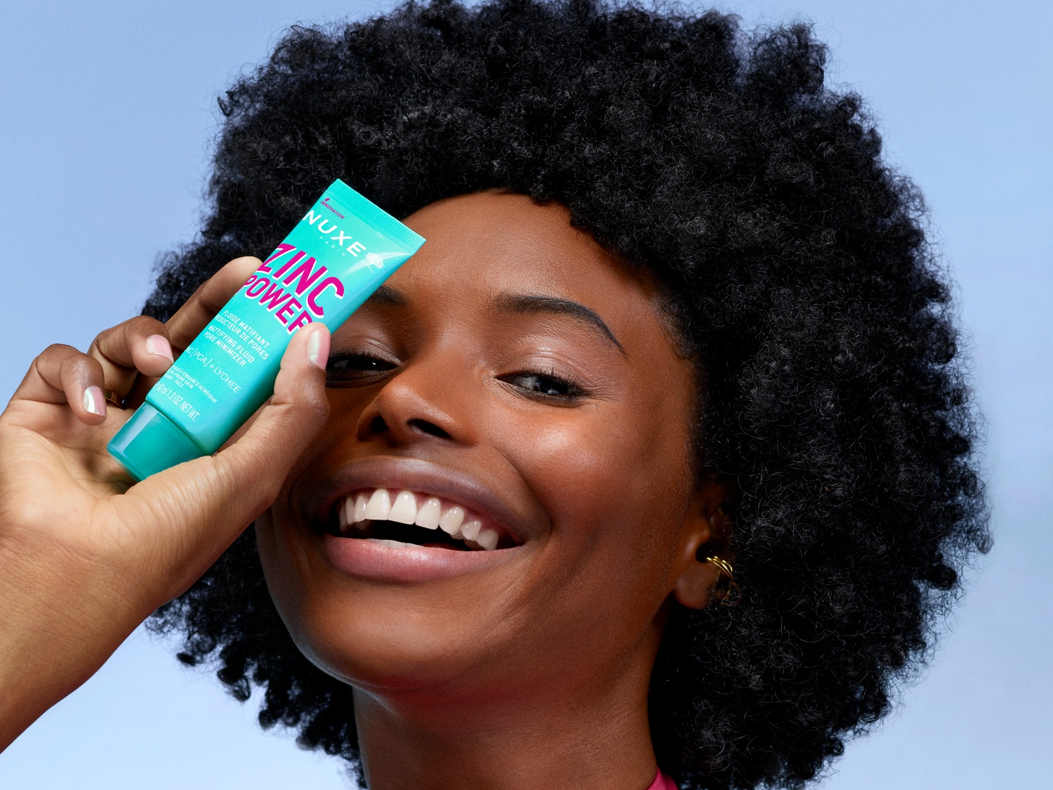 Une femme souriante tient un tube bleu turquoise de Nuxe Zinc Power, un fluide matifiant qui réduit les pores et contient du zinc PCA et du litchi.