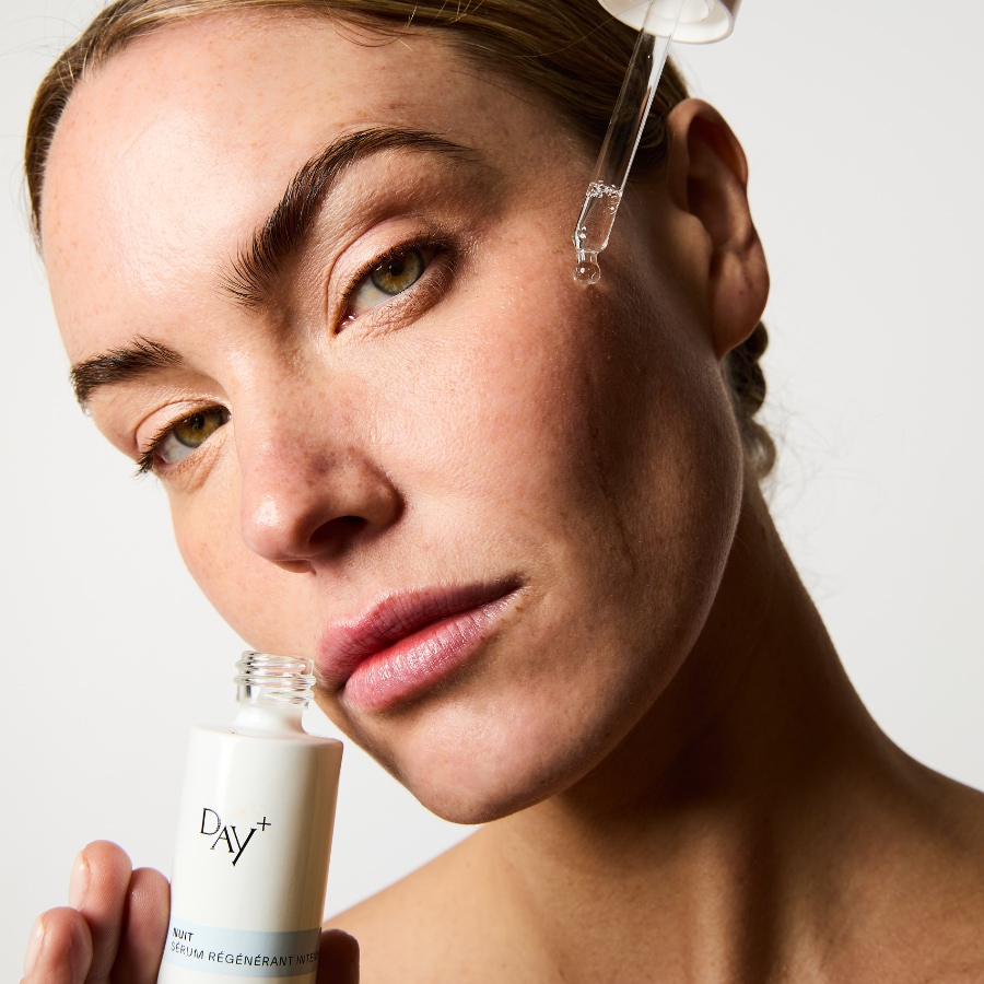Une femme applique le sérum régénérant intense Day+ Nuit sur sa joue à l'aide d'un compte-gouttes, le flacon blanc et bleu est visible en bas à gauche.
