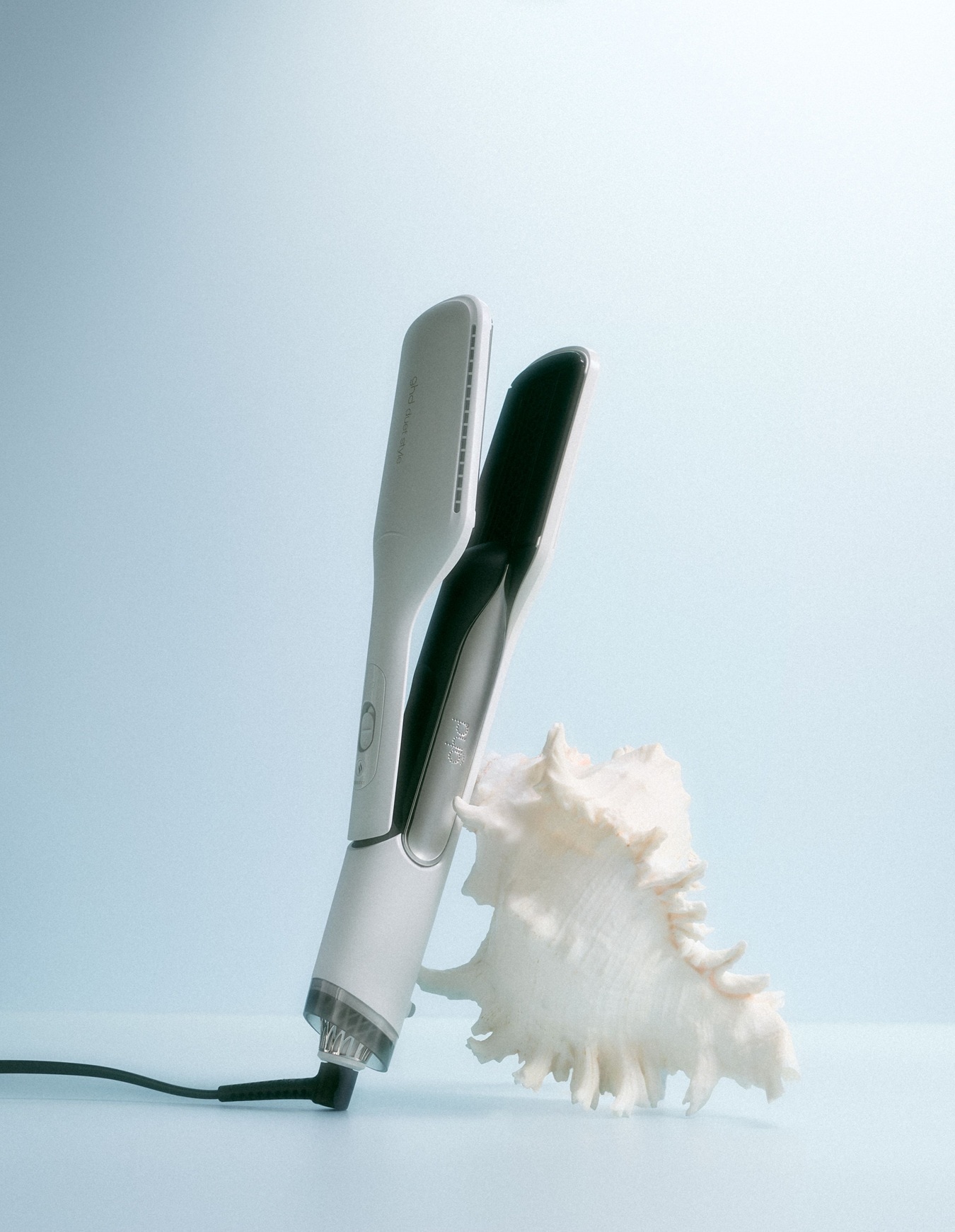 Fer à lisser blanc et noir élégant de GHD, posé à côté d'un coquillage blanc sur un fond bleu clair, pour un coiffage parfait.