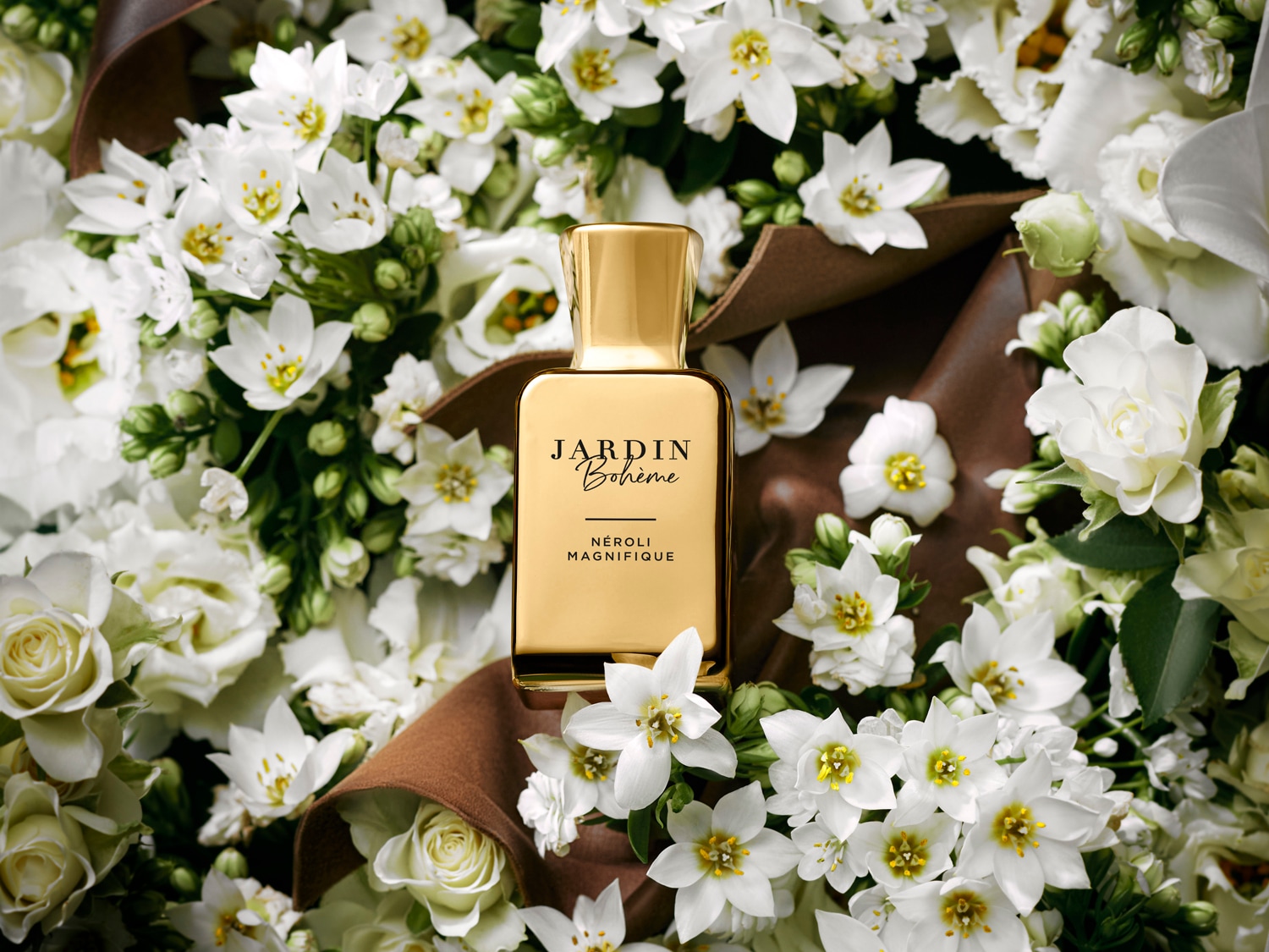 Auksinis „Jardin Bohème Neroli Magnifique“ buteliukas apsuptas baltų gėlių, suteikiantis prabangos ir gaivos įspūdį.