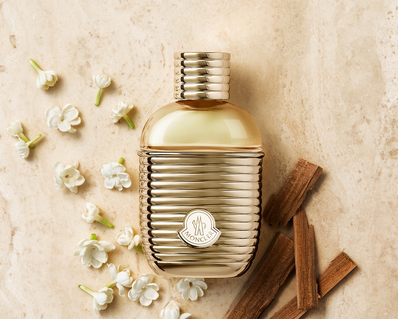 Ein goldener Moncler-Parfümflakon liegt auf einem beigen Untergrund, umgeben von kleinen weißen Blüten und Holzstücken.