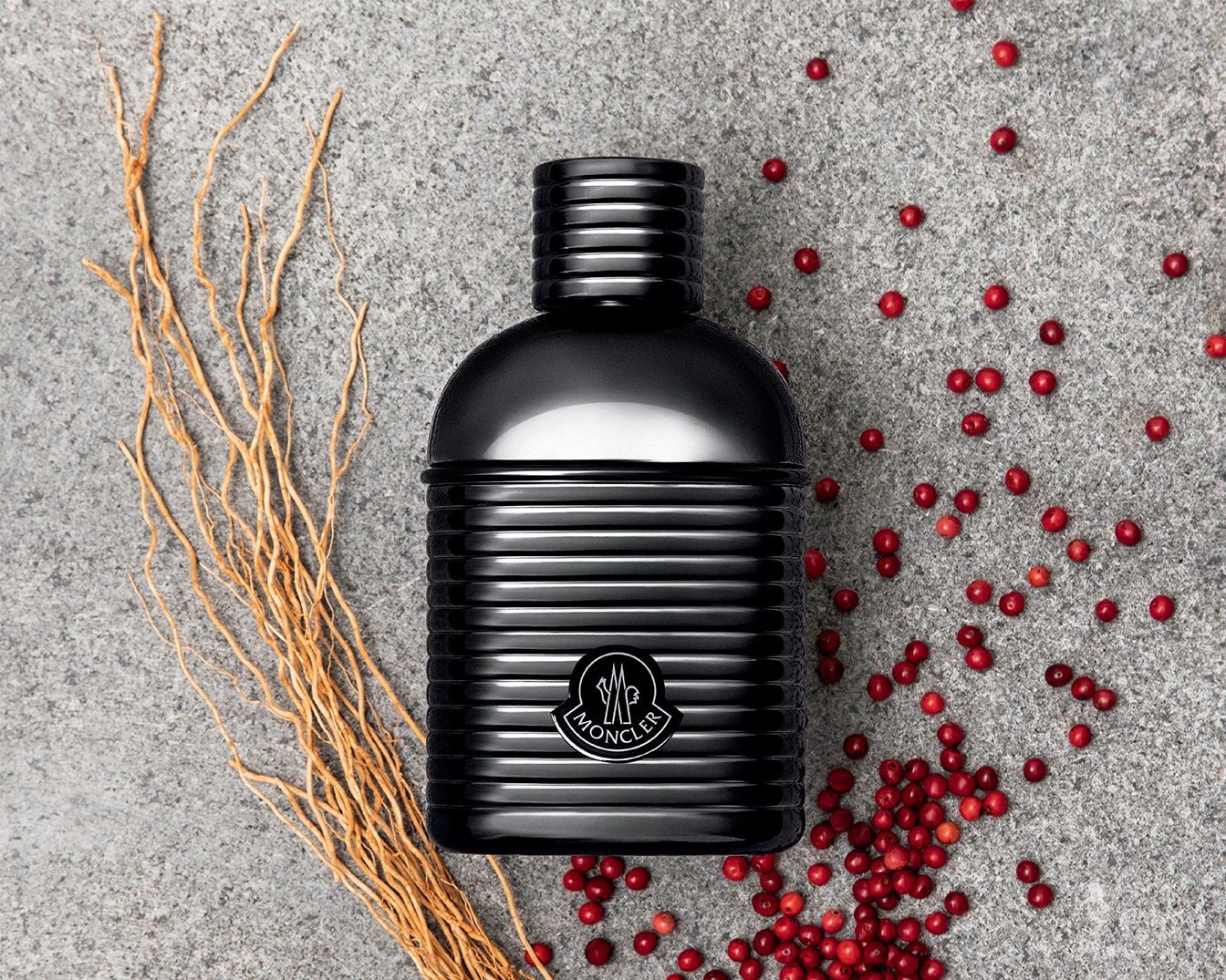 Schwarzer Moncler-Parfümflakon mit geripptem Design, umgeben von roten Beeren und einem Bündel trockener Zweige auf einem grauen Hintergrund.