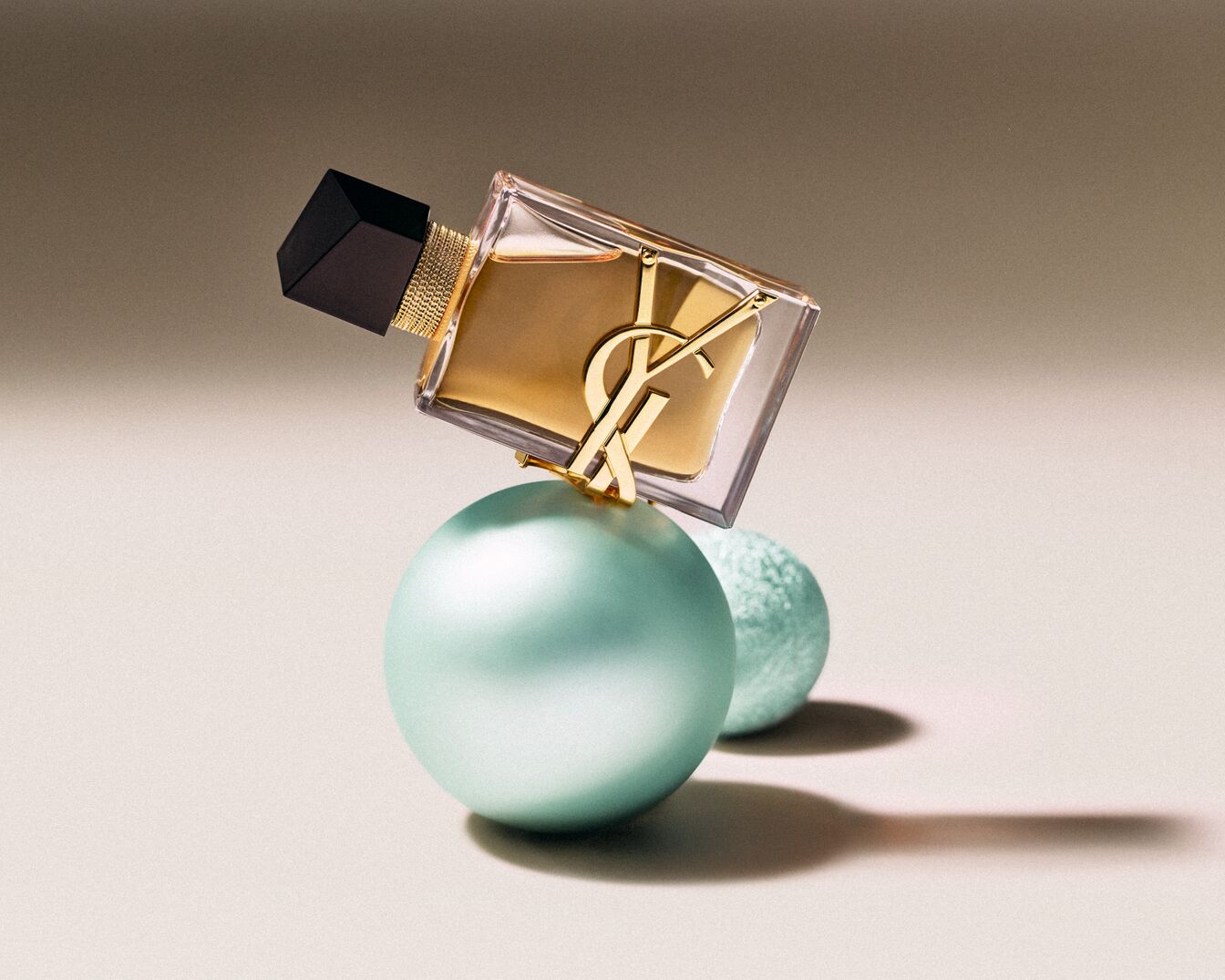 Az YSL Libre Eau de Parfum átlátszó üvegben, arany színű folyadékkal, fekete kupakkal és arany színű YSL logóval, egy nagy és egy kis kék gömbön egyensúlyozva.