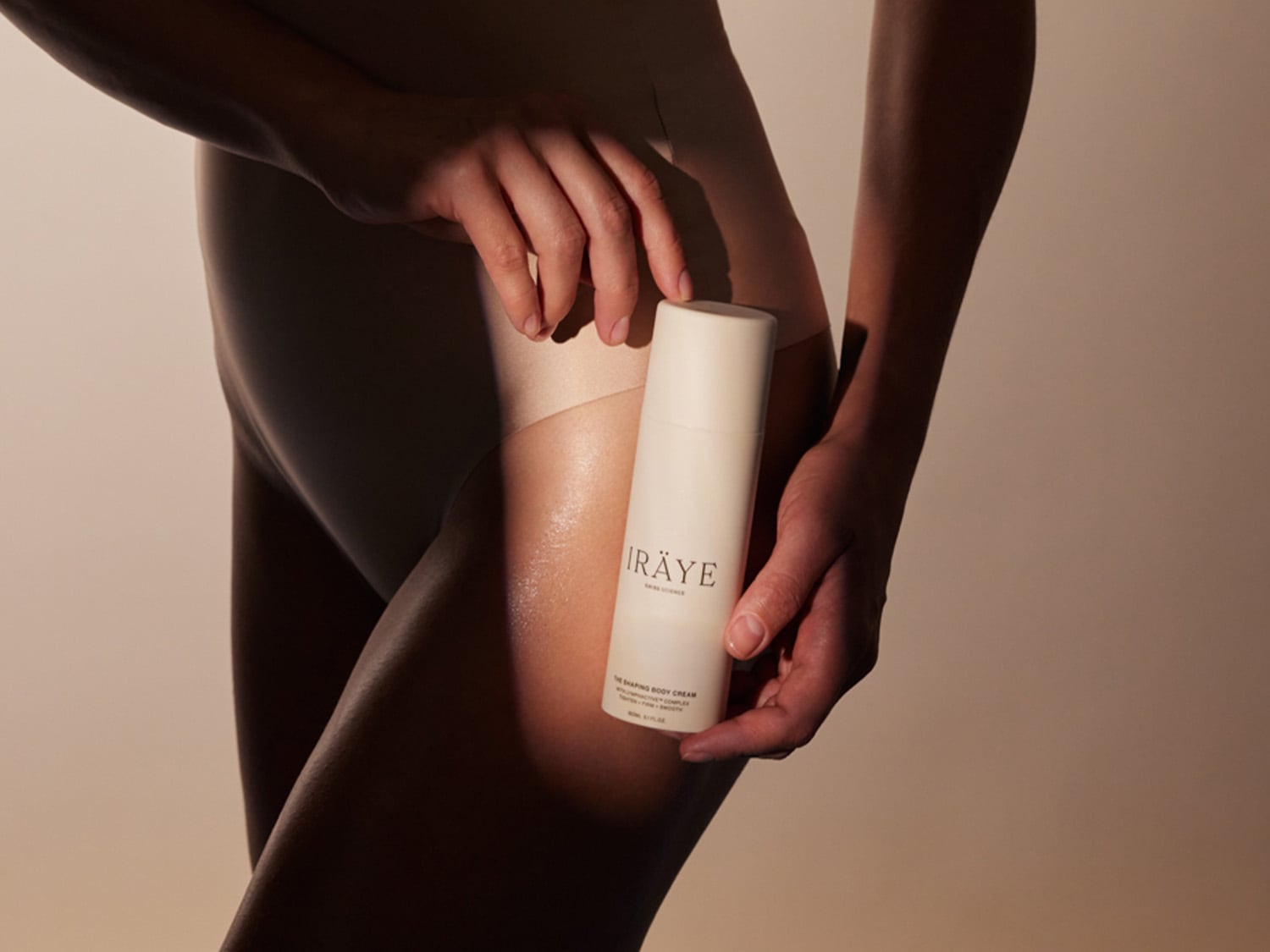 Eine Person in hautfarbenen Body hält eine weiße Tube IRÄYE The Shaping Body Cream auf Höhe ihrer Hüfte.