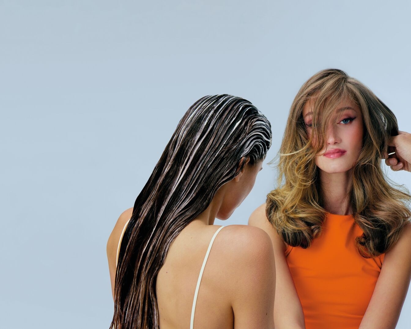 Eine Frau mit dunklem, nassem Haar, auf das eine helle Spülung aufgetragen ist, steht neben einer Frau mit blondem, gewelltem Haar und einem orangefarbenen Oberteil.