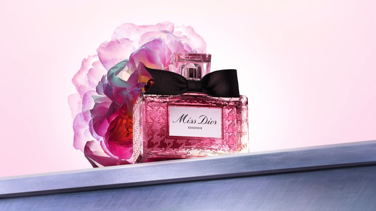 Ein rosa Miss Dior Essence Parfümflakon mit einer schwarzen Schleife steht vor einer verschwommenen rosa Blume.