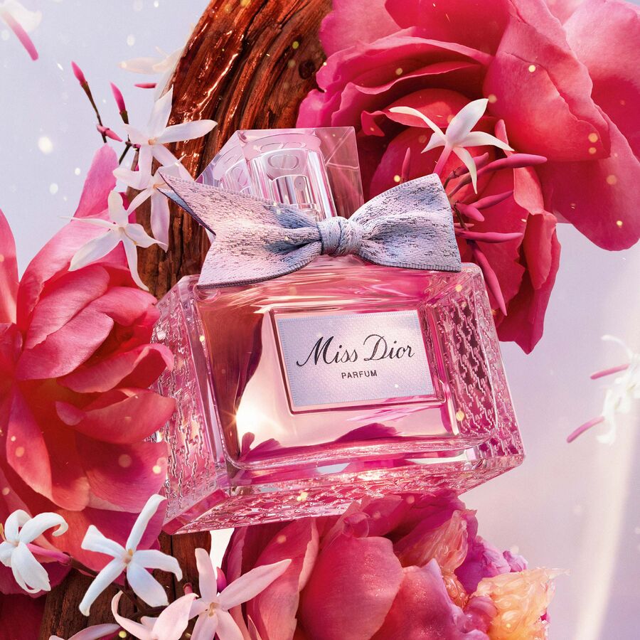 Ein rosafarbener Miss Dior Parfümflakon mit einer Schleife sitzt inmitten von rosa Blüten und kleinen weißen Blüten.