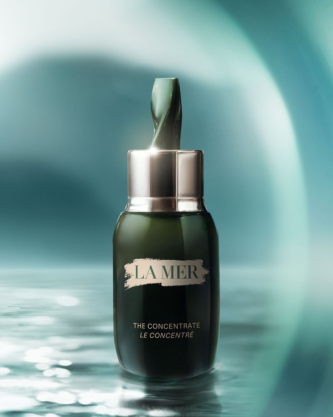 De La Mer 'The Concentrate' wordt gepresenteerd in een donkergroene fles met zilveren dop, met een glanzende textuur die de essentie van de zee weerspiegelt.