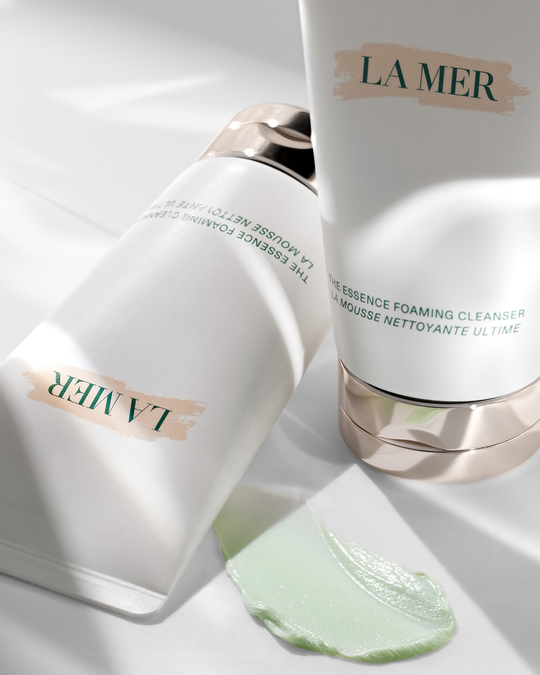 Twee witte tubes La Mer The Essence Foaming Cleanser liggen op een witte ondergrond, met een vleugje lichtgroene reiniger ernaast.