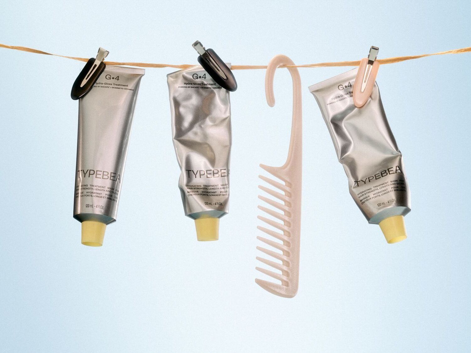 Trys „TYPEBEA“ plaukų priežiūros priemonės sidabriniuose tūbelėse su geltonais dangteliais, pakabintos ant virvės su drabužių segtukais, šalia šviesiai smėlio spalvos šukų.