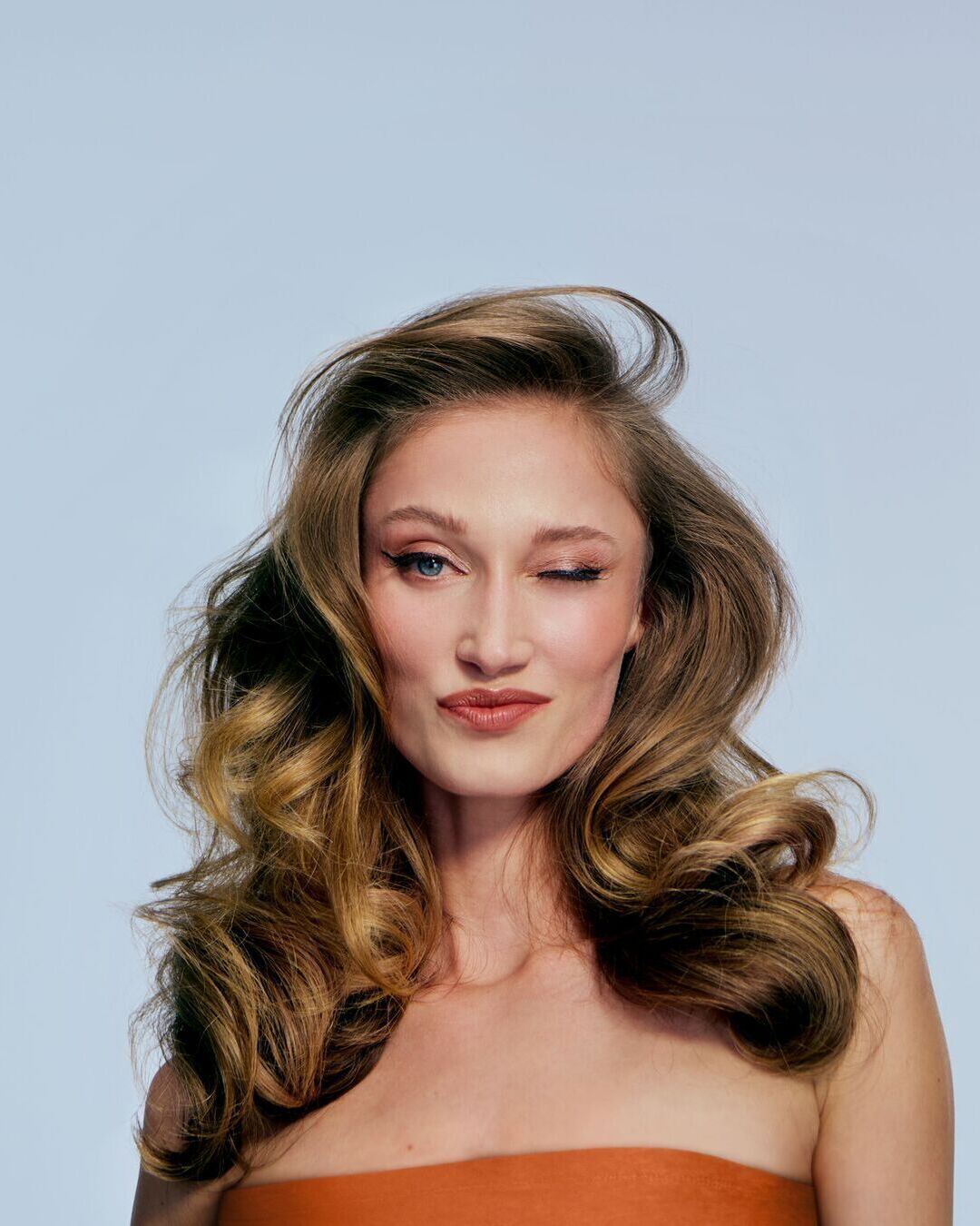 Šviesiaplaukė moteris su šviesiai mėlynomis akimis mirksi į kamerą, jos plaukai garbanoti ir krenta ant pečių.