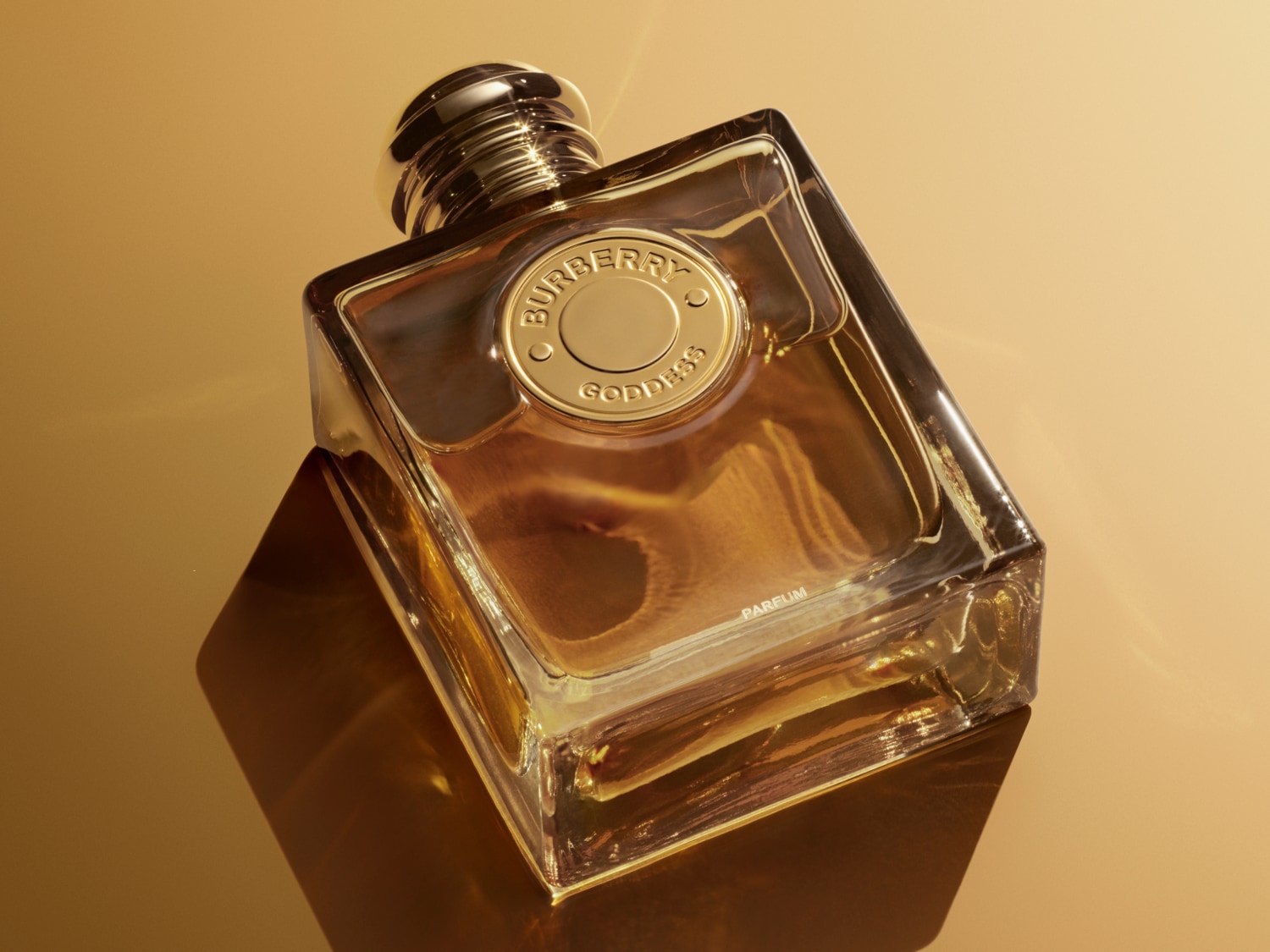 Butelka perfum Burberry Goddess o kwadratowym kształcie z przezroczystego szkła z płynem w kolorze bursztynowym, złotą nakrętką i logo marki na tle krajobrazu.