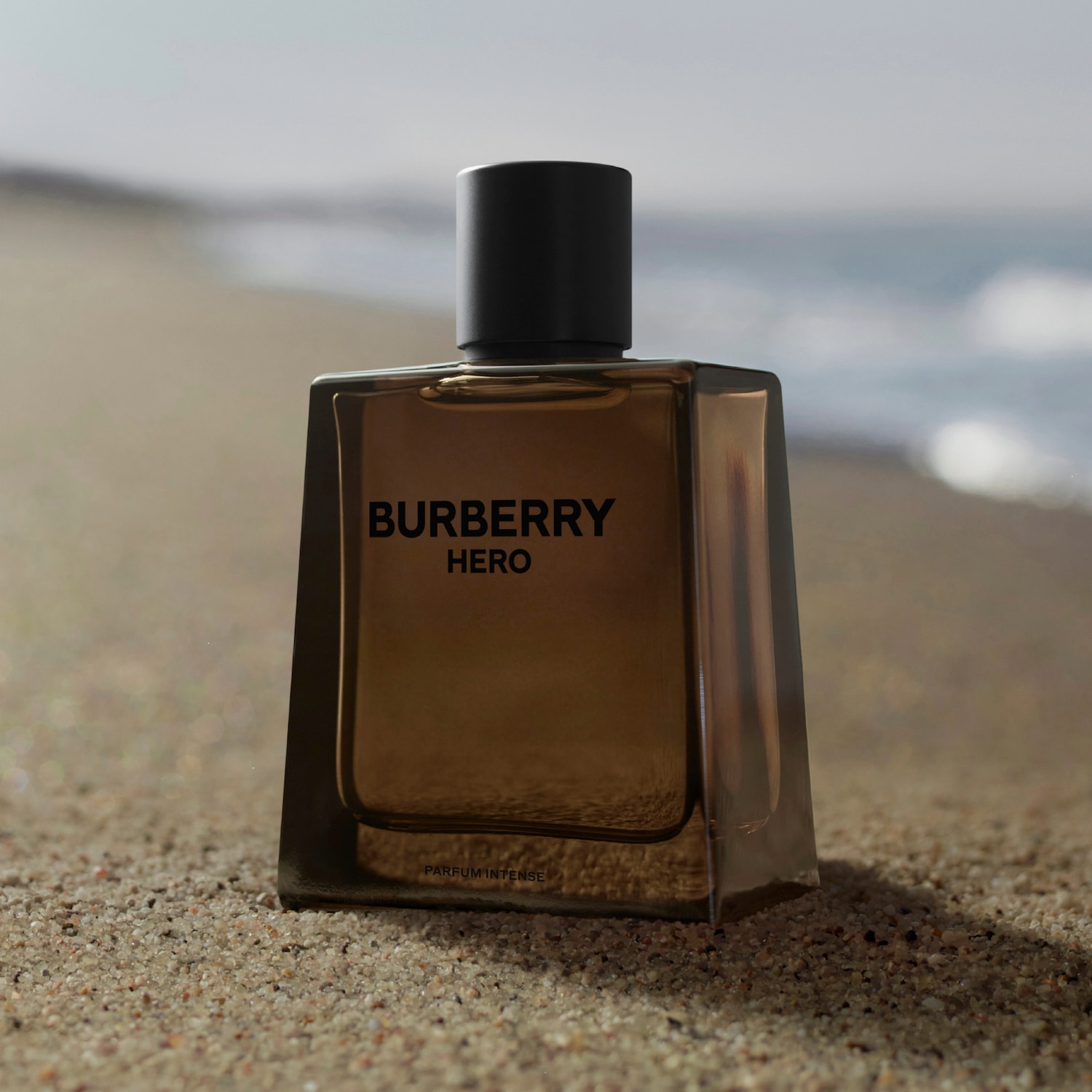 Butelka brązowej wody perfumowanej Burberry Hero z czarną zakrętką stoi na mokrej powierzchni, obok niej leżą kawałki drewna i zielona roślina.