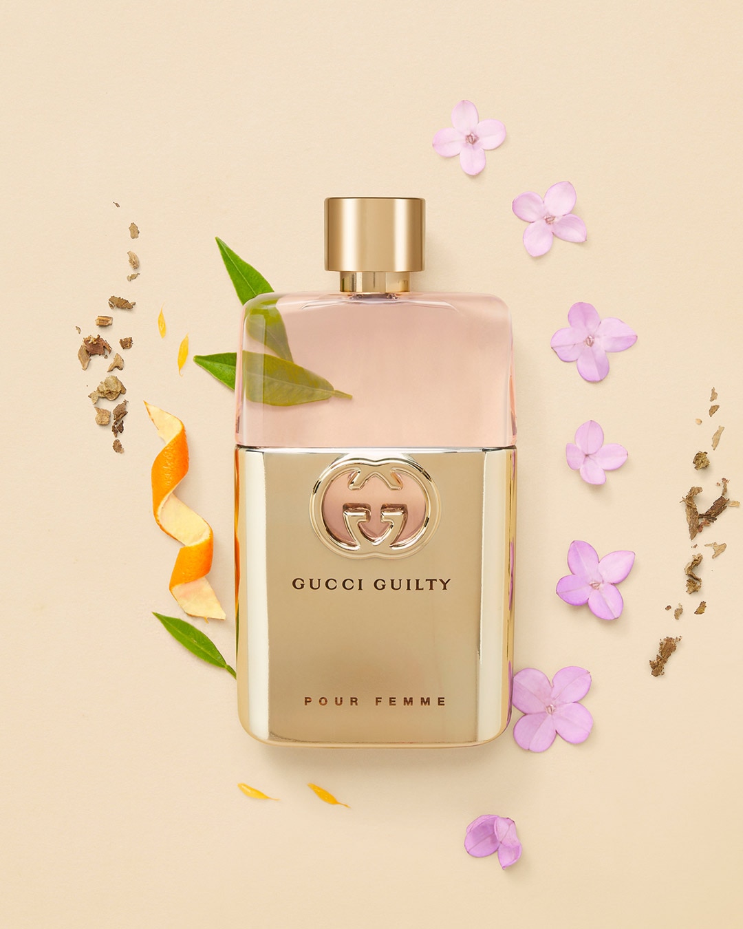 Butelka perfum Gucci Guilty Pour Femme w złotym flakonie, otoczona kwiatami i skórką pomarańczy, tworzy elegancką kompozycję.