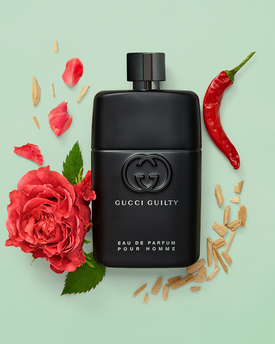 Czarny flakon perfum Gucci Guilty Eau de Parfum pour Homme otoczony czerwoną różą, papryczką chili i drewnianymi wiórkami na jasnym tle.