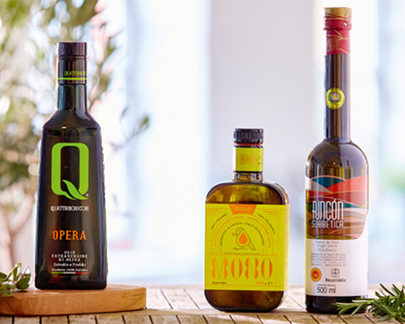 Drei Flaschen Olivenöl unterschiedlicher Marken stehen auf einem Holztisch, jede mit einzigartigen Etiketten und Formen.