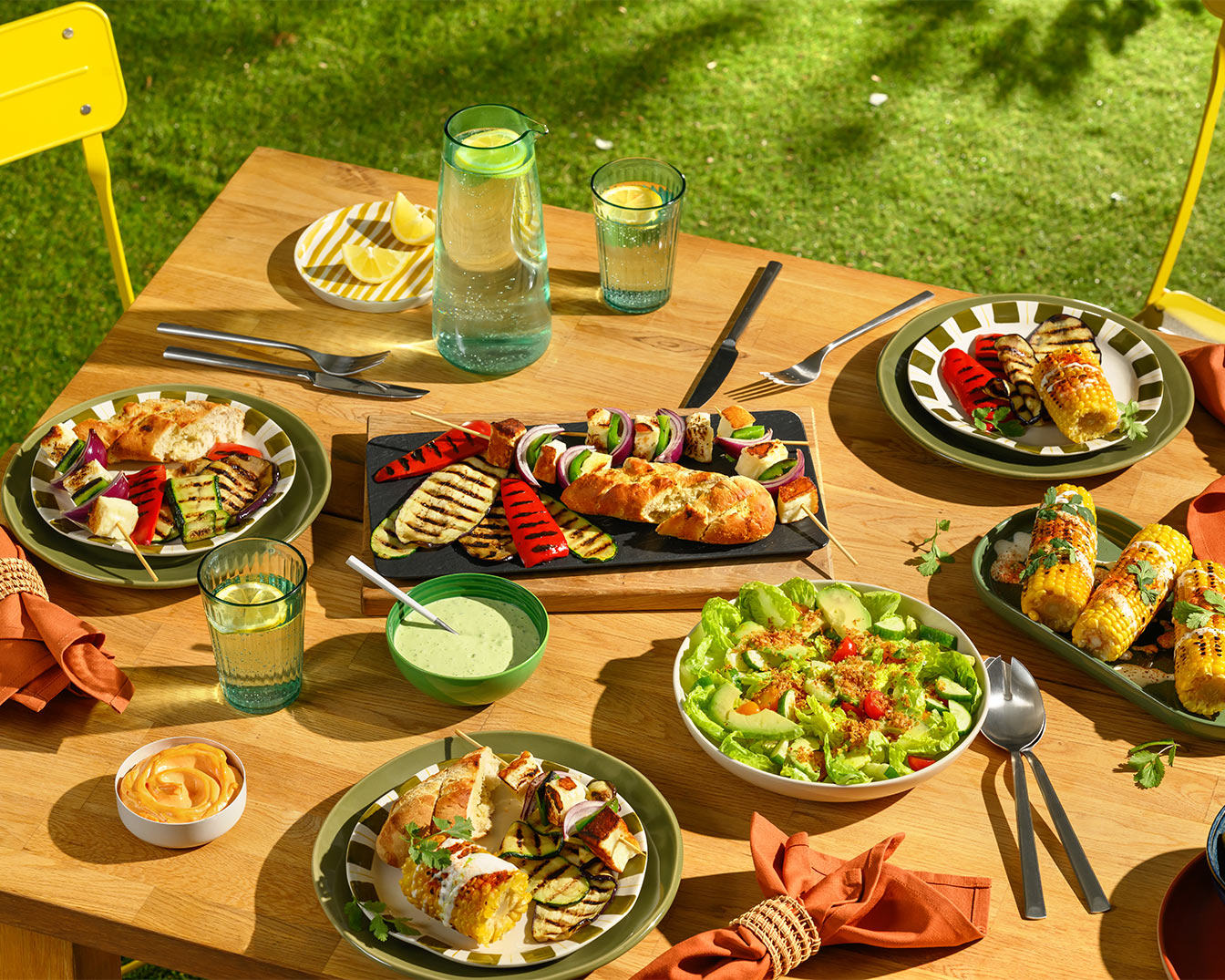 Draufsicht auf einen Holztisch mit gegrilltem Gemüse, Maiskolben, Salat und Getränken, arrangiert für ein sommerliches Essen im Freien.