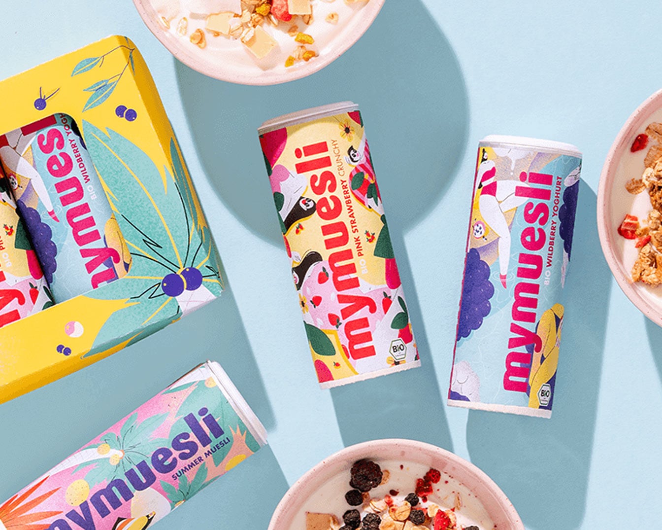 Drei Mymuesli-Dosen in verschiedenen Geschmacksrichtungen, darunter Sommer Müsli, rosa Erdbeer-Crunchy und Wild Berry Joghurt, zusammen mit einer gelben Müsli-Box und Müslischalen.
