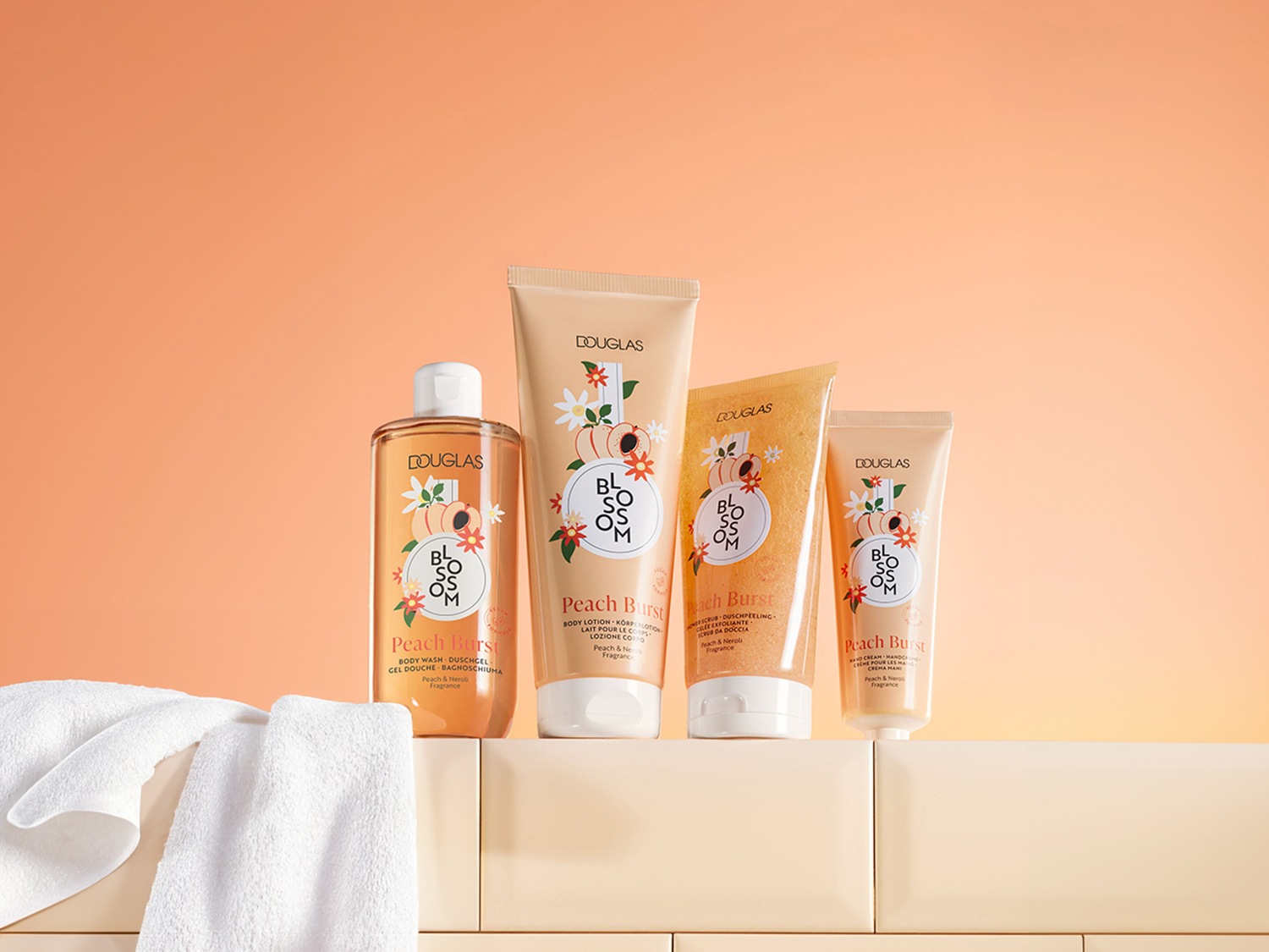 Vier Douglas Blossom Peach Burst-producten staan op een rij, waaronder body wash in een heldere fles, bodylotion, bodyscrub en handcrème in beige tubes, allemaal met perzik- en neroli-geur.