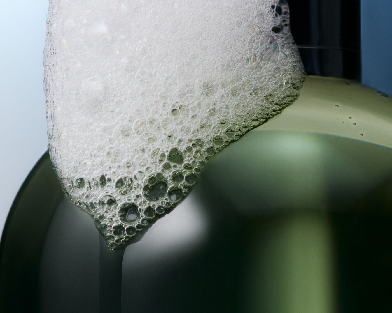Gros plan sur un flacon vert translucide coiffé d'une mousse blanche et aérée, évoquant la fraîcheur et la pureté d'un soin nettoyant.