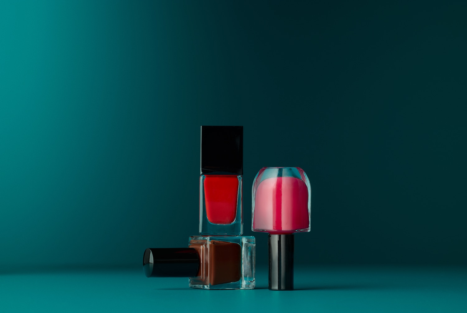 Découvrez un trio de vernis à ongles : un rouge vif en flacon carré, un rose doux en flacon arrondi, et un brun riche en flacon carré, tous sur un fond turquoise.