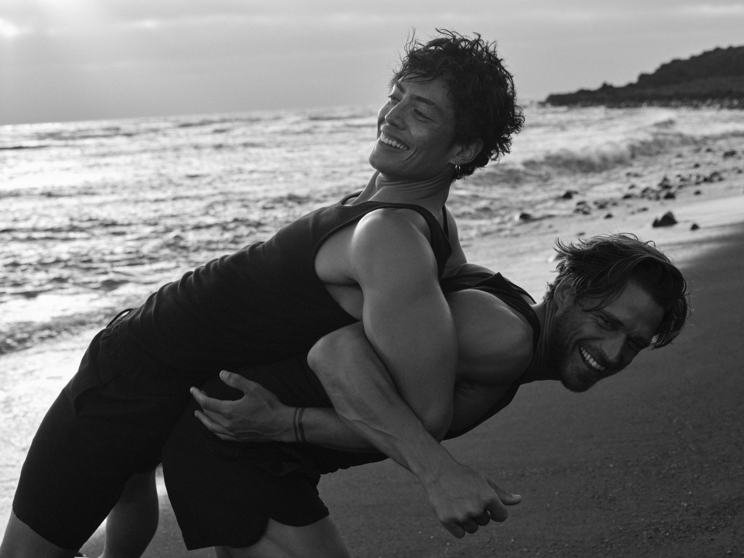 Schwarz-Weiß-Foto von zwei Männern am Strand, einer trägt den anderen Huckepack, beide lächeln und genießen die Zeit.