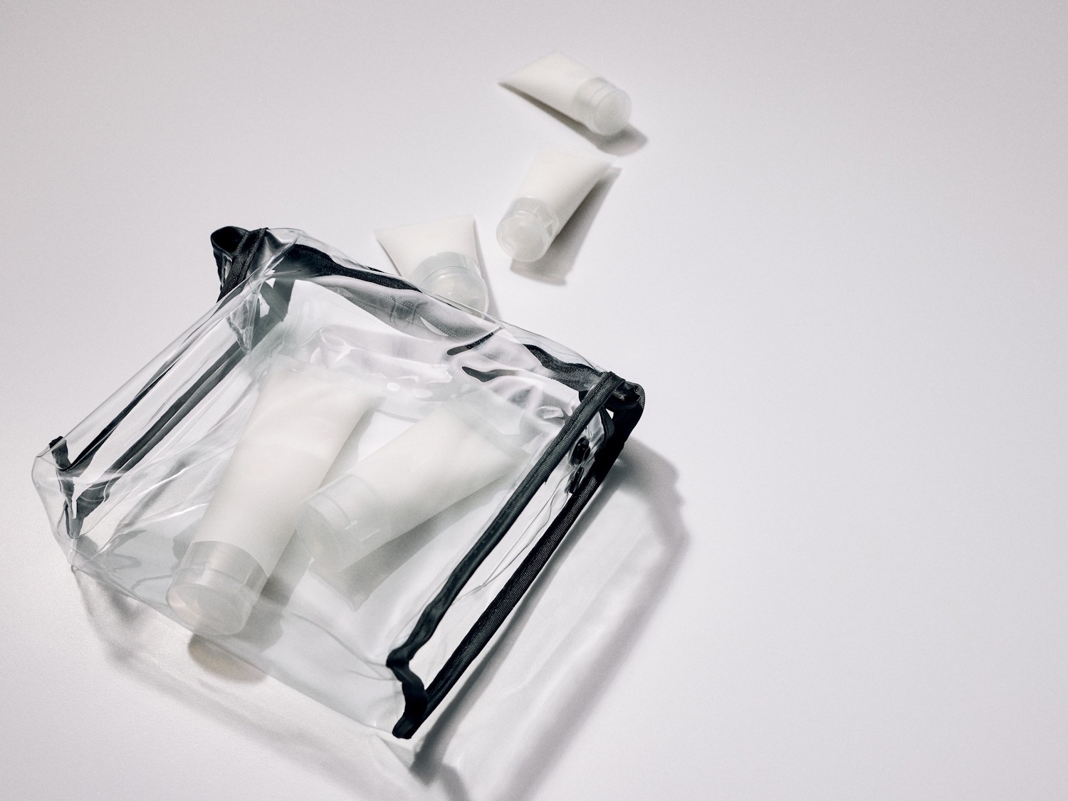 Átlátszó, fekete szegélyű kozmetikai táska, benne fehér, utazáshoz való minitubusokkal, néhány tubus pedig a táska mellett helyezkedik el.