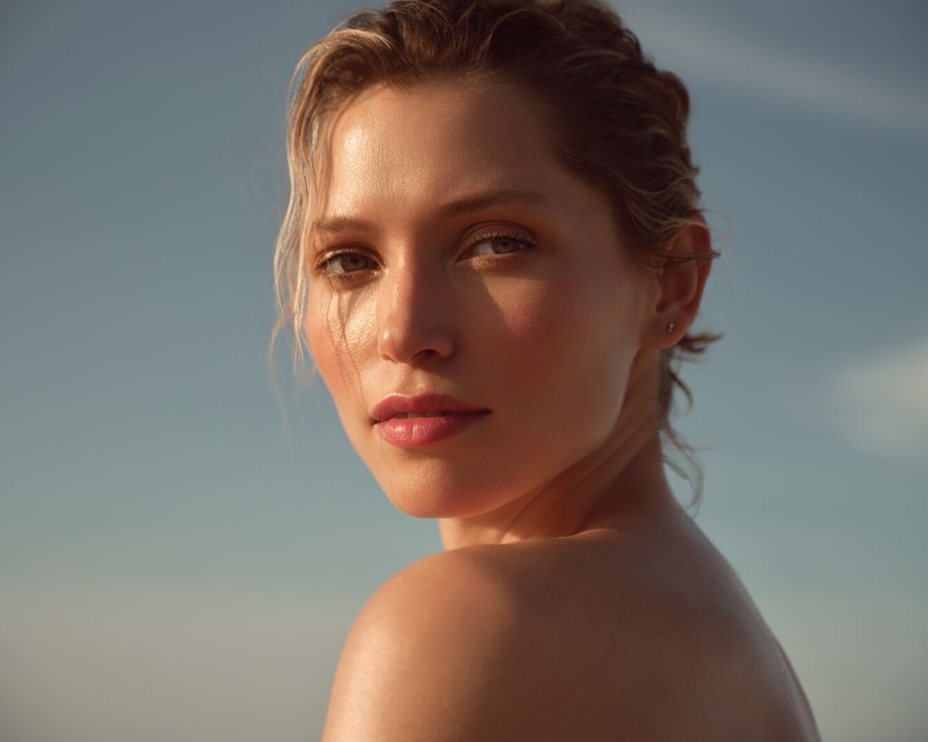 A képen egy világos bőrű, szőke hajú nő látható, aki a kamerába néz, enyhe sminkkel, ami kiemeli a természetes szépségét.