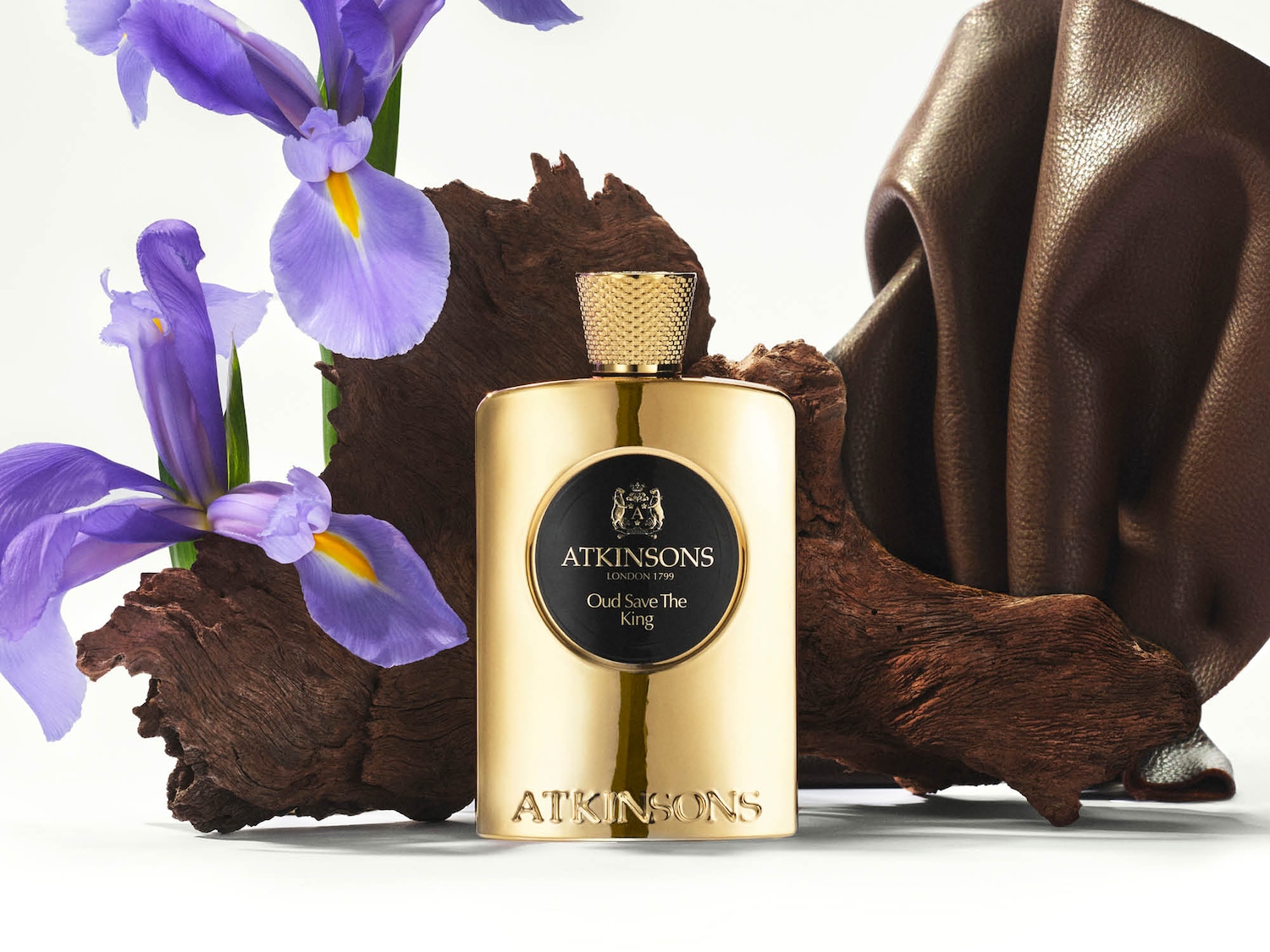 Goldener Flakon von Atkinsons Oud Save The King, umgeben von lila Irisblüten, einem Stück Holz und drapiertem braunem Leder.