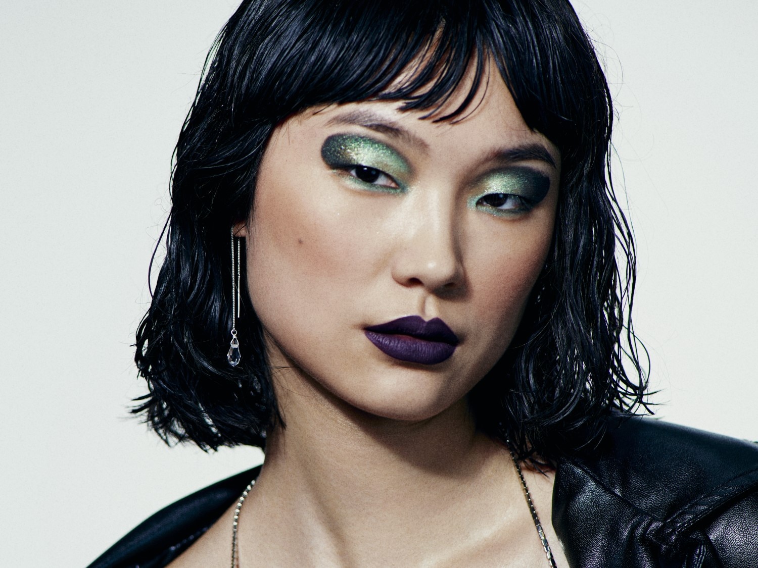 Nahaufnahme eines asiatischen Models mit glänzendem, grünem Lidschatten, dunklem Lippenstift und nassen, schwarzen Haaren, das eine silberne Kette und Ohrringe mit Tropfenanhänger trägt.
