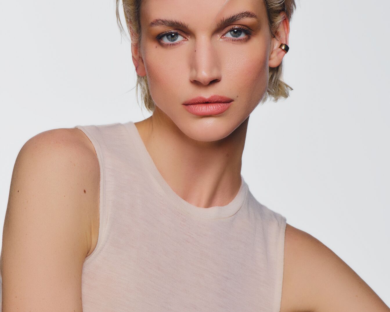 Porträt einer Frau mit blonden, kurzen Haaren, hellblauen Augen und einem goldenen Ohrring, die ein helles, ärmelloses Oberteil trägt.