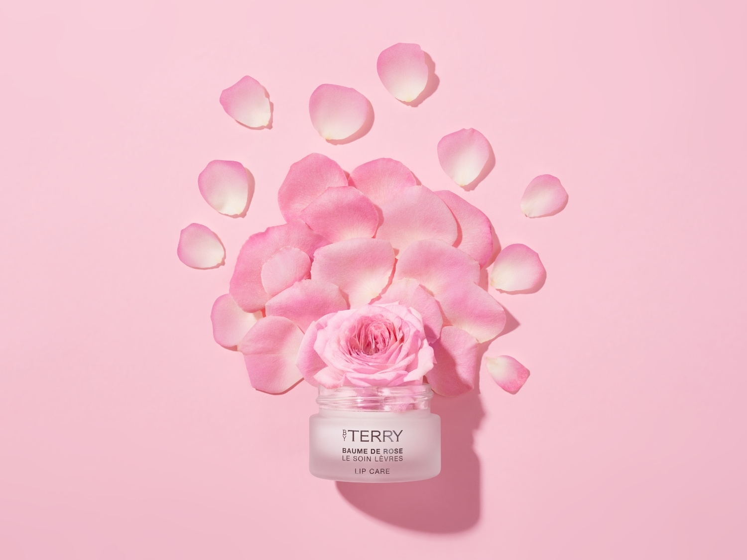 Na różowym tle leży słoiczek balsamu do ust By Terry Baume de Rose Lip Care, otoczony płatkami róż i różą, tworząc delikatną kompozycję.