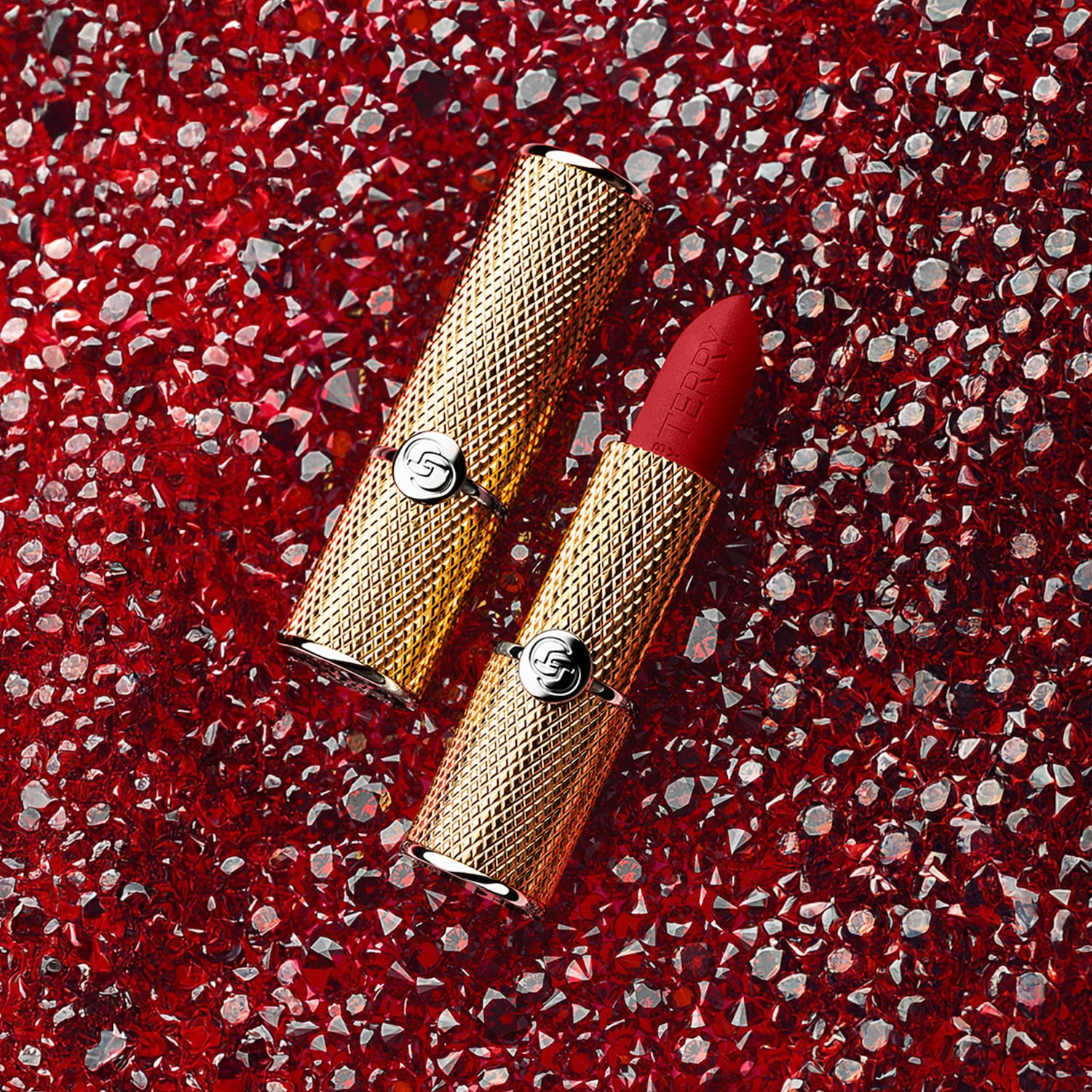Szminka Terry w złotym opakowaniu na tle czerwonych kryształków, oferująca luksusowy akcent kolorystyczny.