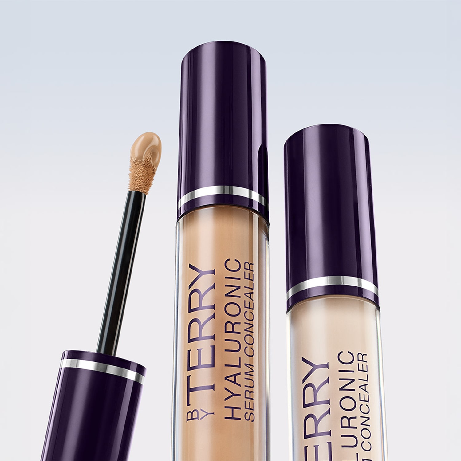 Dwa korektory do twarzy By Terry Hyaluronic Serum Concealer w odcieniach beżu i ecru, z ciemnofioletowymi zakrętkami i aplikatorami.