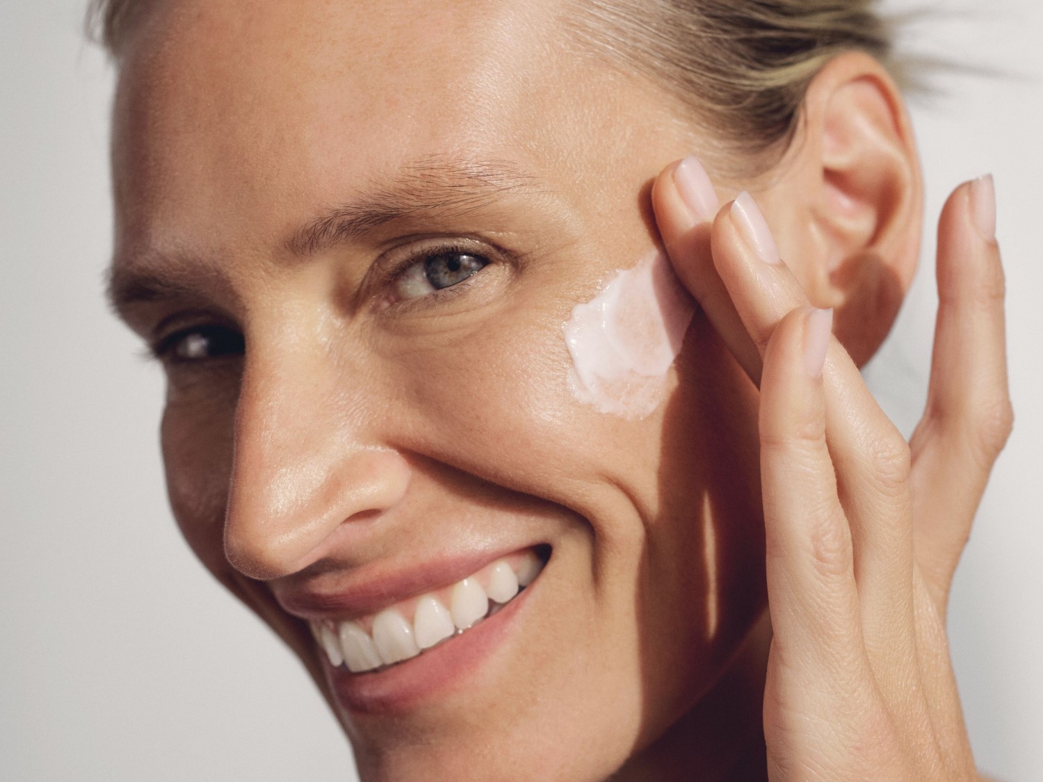 Een vrouw lacht terwijl ze een witte crème op haar gezicht aanbrengt met haar vingers, wat zorgt voor een stralende en gehydrateerde huid.