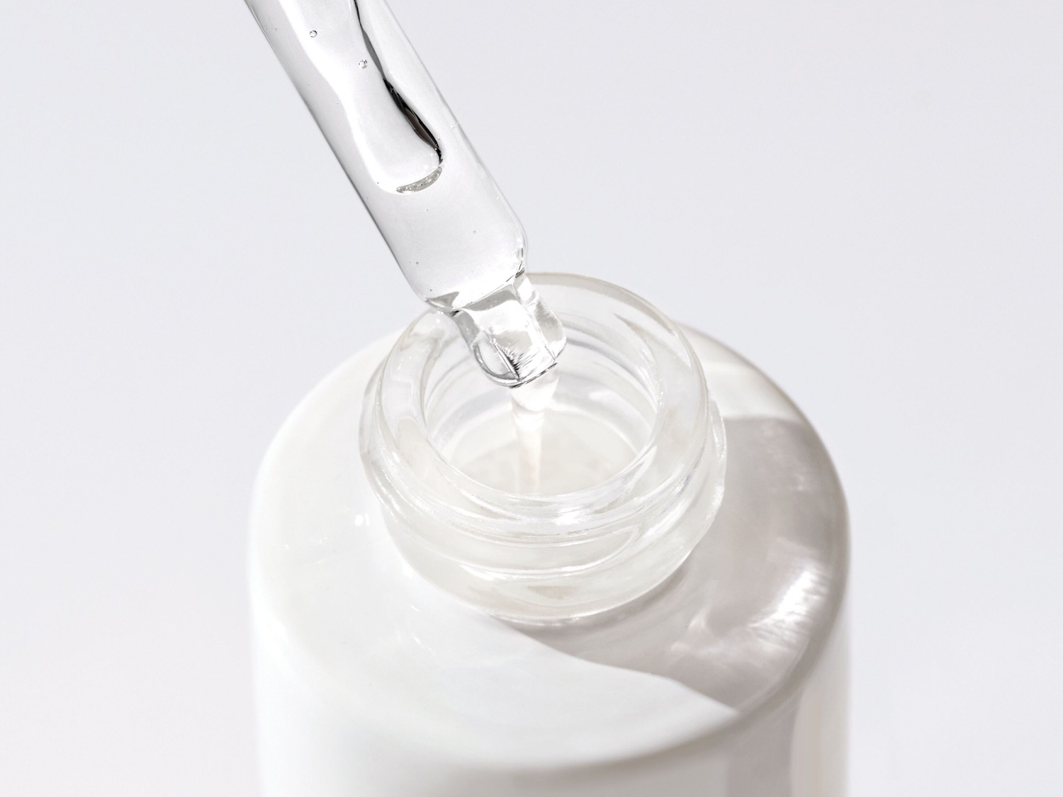 Een heldere vloeistof druppelt uit een glazen pipet in een witte fles, wat de pure essentie van het serum benadrukt.