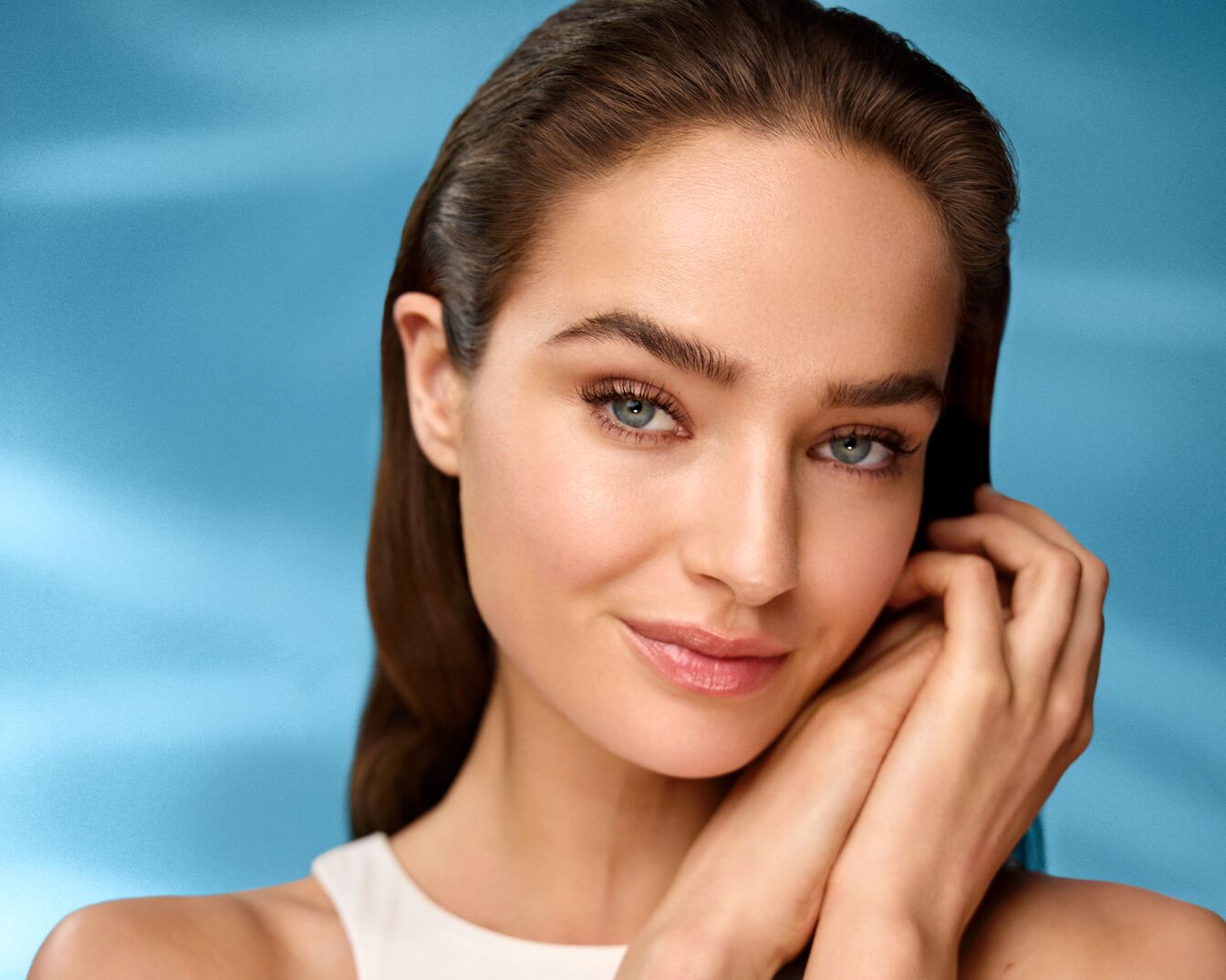 A képen egy fiatal nő látható, sima, világos bőrrel, enyhe mosollyal, és a kezét az arcához emeli, kiemelve természetes szépségét.