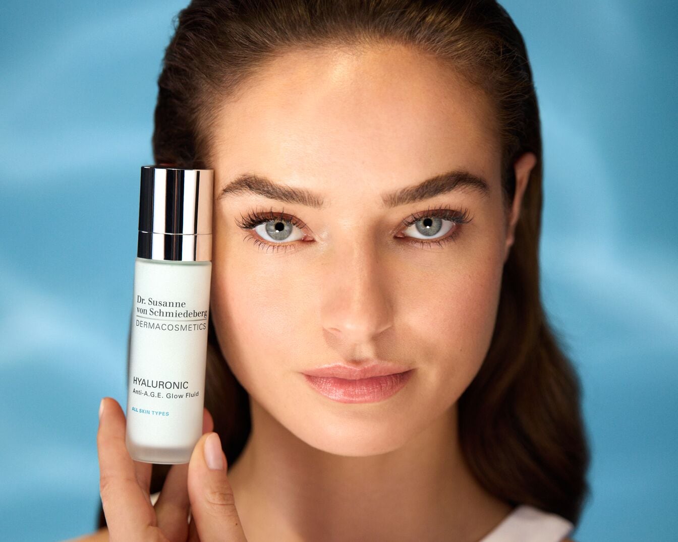 A képen egy nő látható, aki a Dr. Susanne von Schmiedeberg DERMACOSMETICS Hyaluronic Anti-A.G.E. Glow Fluid termékét tartja, mely minden bőrtípusra alkalmas.