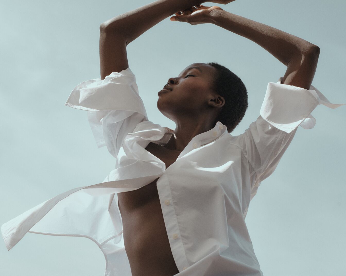 Egy fekete bőrű nő laza, fehér inget visel, karjait a feje fölé emeli, és élvezi a napsütést.