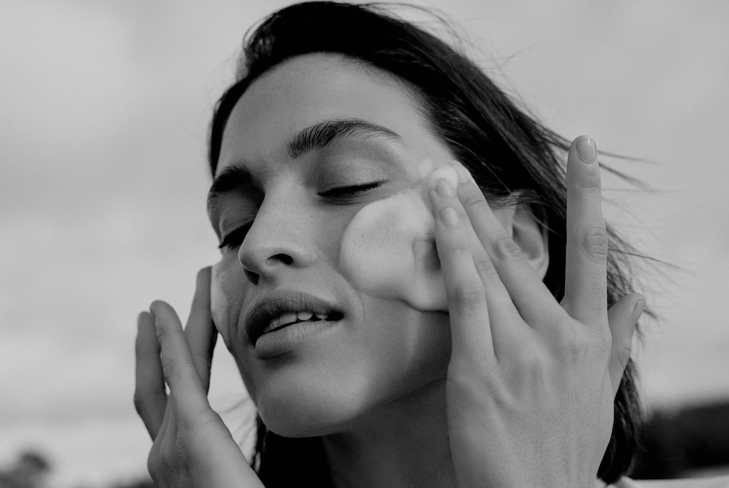 Eine Frau mit geschlossenen Augen massiert ihr Gesicht mit einer schaumigen Substanz, um ihre Haut zu reinigen und zu pflegen.