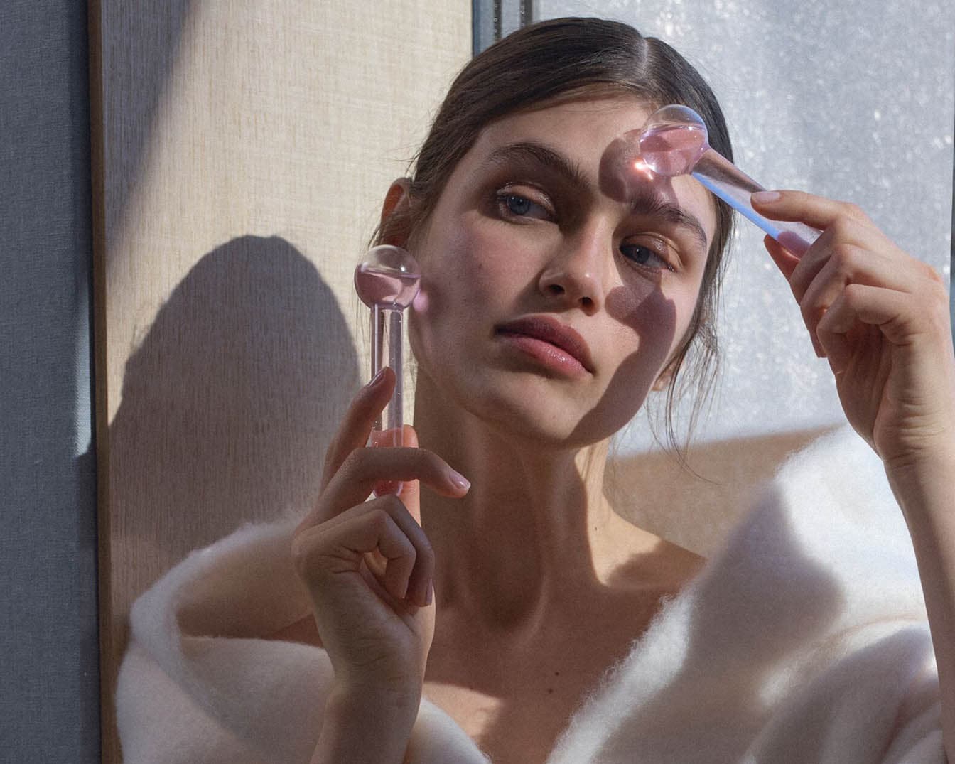 Eine Frau mit blauen Augen benutzt zwei rosafarbene Kühlkugeln auf ihrem Gesicht, um ihre Haut zu erfrischen und zu beruhigen.