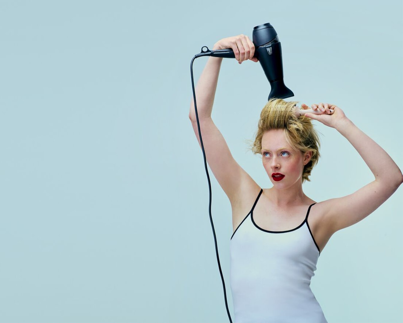 Een vrouw met een wit hemdje föhnt haar blonde haar met een zwarte föhn tegen een lichtblauwe achtergrond.