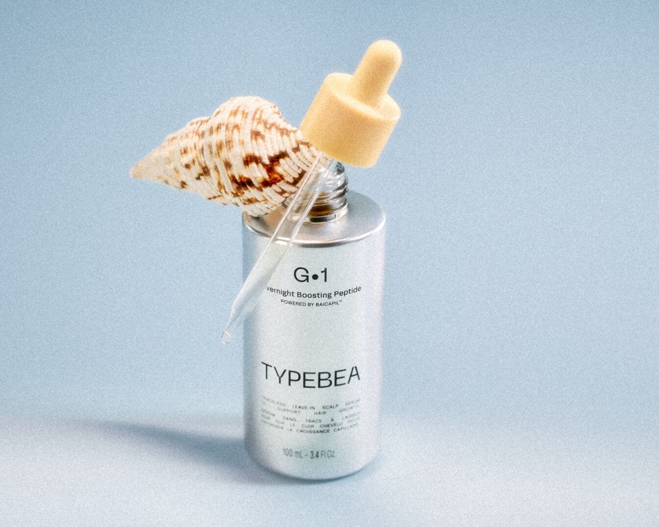 Een zilveren flesje TYPEBEA G.1 Overnight Boosting Peptide serum met een beige dop en een glazen pipet, versierd met een zeeschelp.