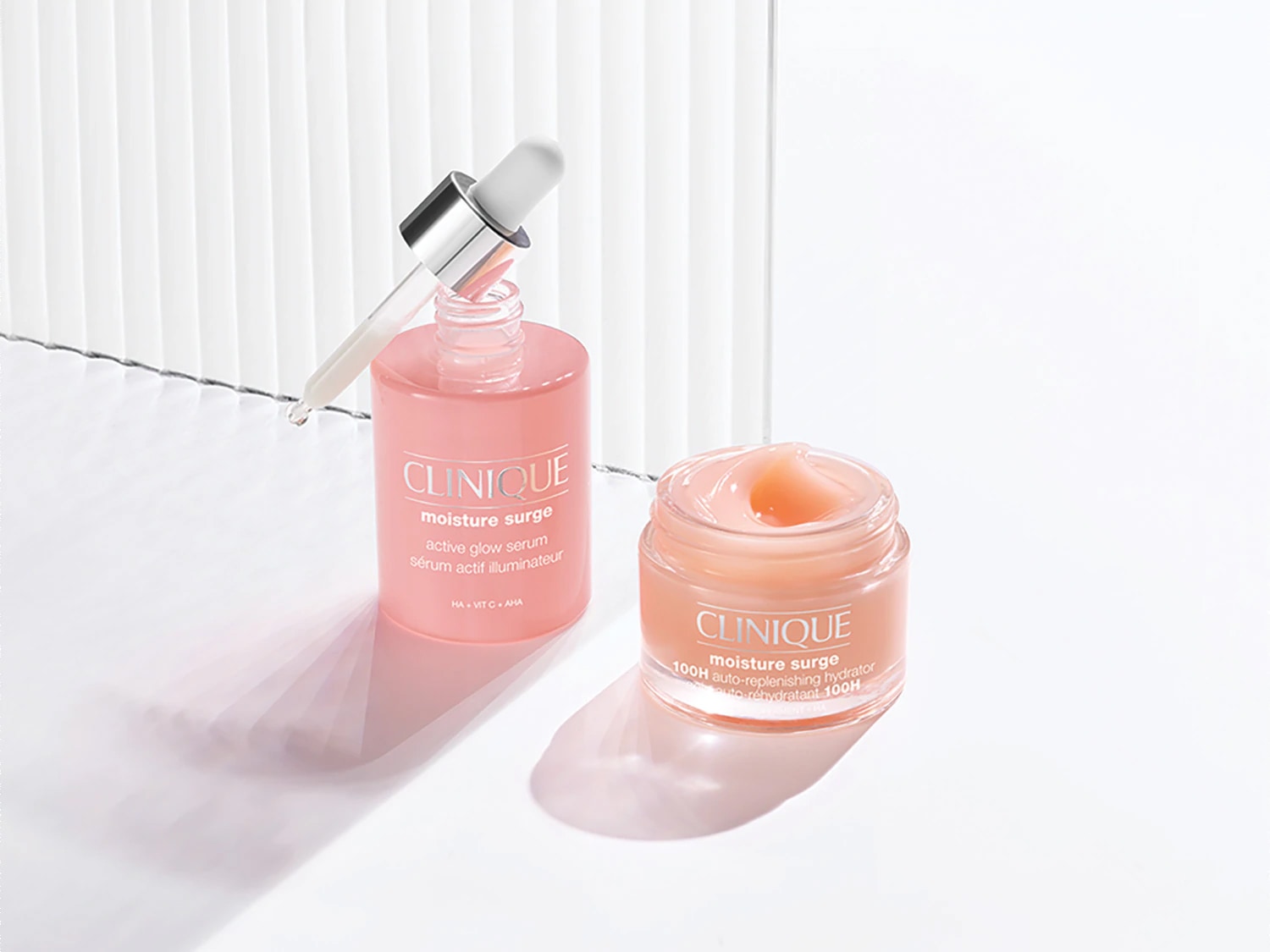 Voici le sérum éclat actif Moisture Surge de Clinique dans un flacon rose avec un compte-gouttes, à côté du hydratant auto-réhydratant 100H Moisture Surge dans un pot transparent.